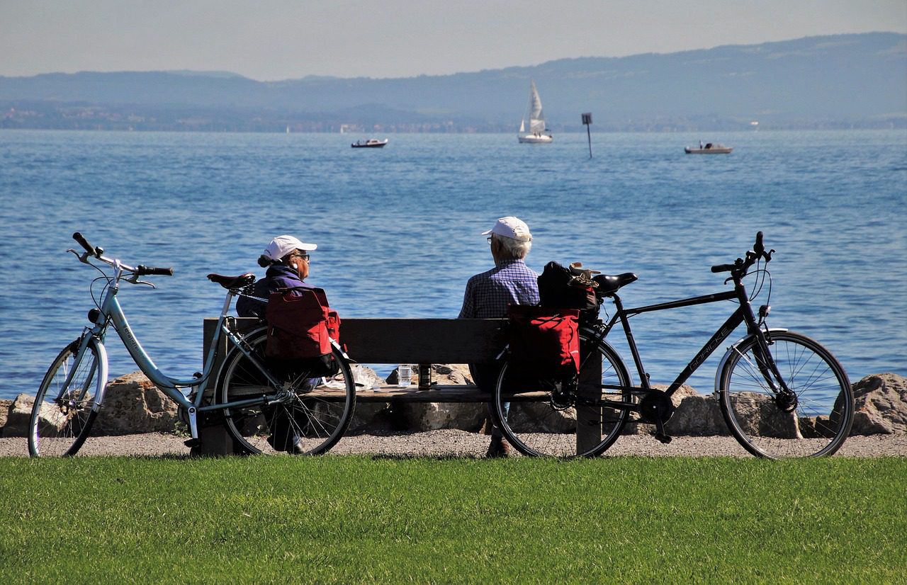 Ouder koppel zit op een bankje aan zee met fietsen naast zich, kijkend naar het water en de bergen, symbool voor rust door geautomatiseerd beleggen.
